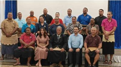 A group of men and women pose together indoors, with most men wearing traditional Tongan attire and some people in business or casual clothes, seated and standing in rows.