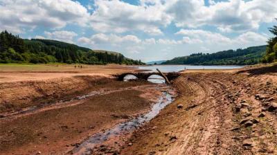 A dried-out riverbed reveals an old stone bridge, surrounded by exposed earth, with a lake and forested hills in the background under a partly cloudy sky.
