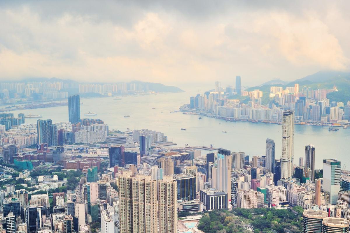 Aerial view of a densely populated city with numerous high-rise buildings by the water, under a cloudy sky.