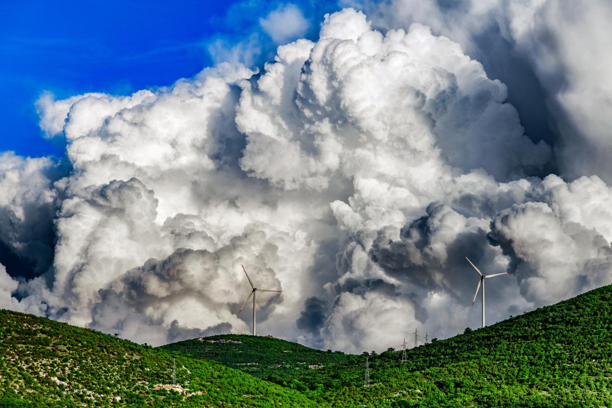 Large, dramatic clouds rise above green hills with wind turbines.