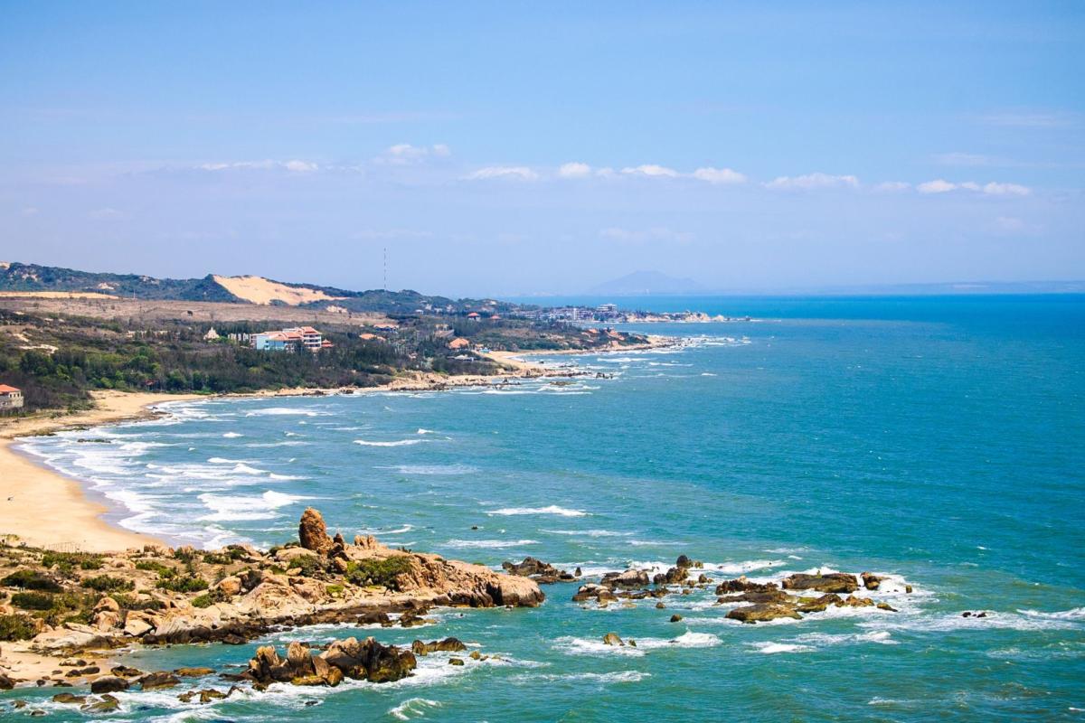 Coastal landscape with sandy beaches, rocky shoreline, and gentle waves under a blue sky, with hills and scattered buildings in the background.