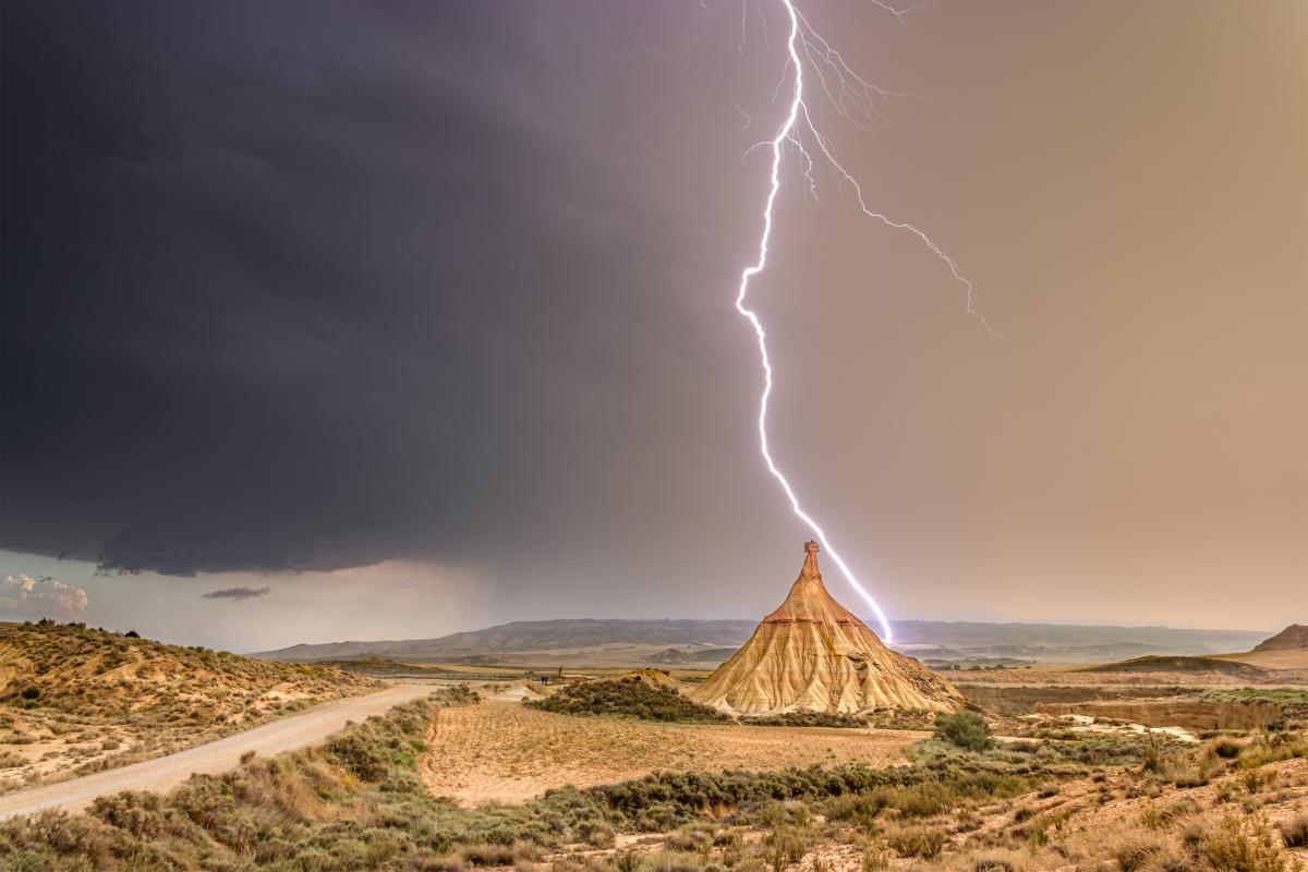 A bolt of lightning strikes a conical rock formation in a desert landscape under a dramatic sky, with a dirt road curving through the scene.