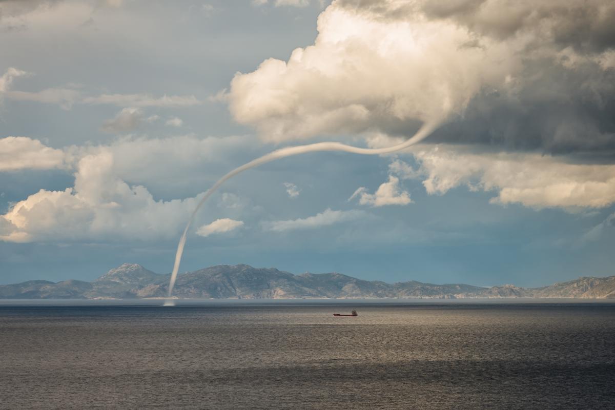 A thin waterspout stretches from a cloudy sky to the sea near a distant mountain range, with a cargo ship visible on the calm water.