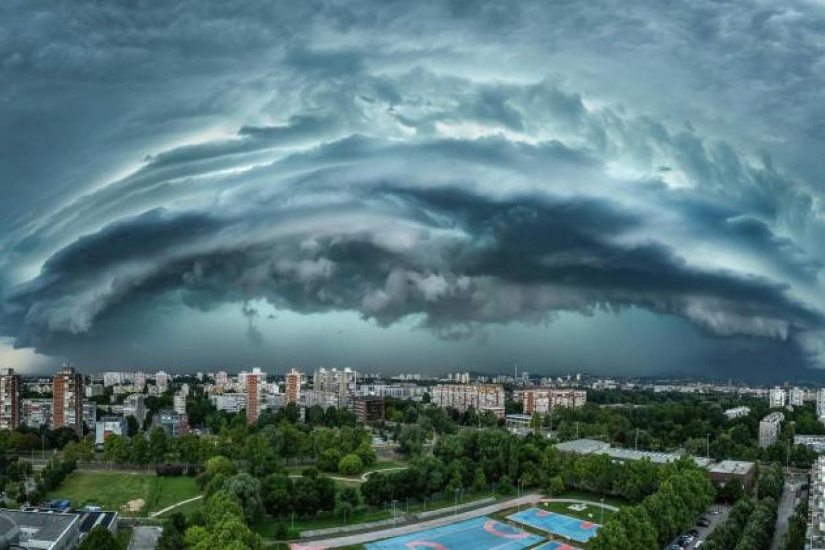 A dramatic, layered storm cloud formation looms over a cityscape with residential buildings and green spaces visible below.