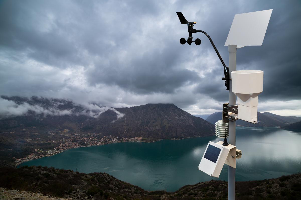 A weather station with sensors and a solar panel stands on a hill overlooking a lake, mountains, and cloudy sky.