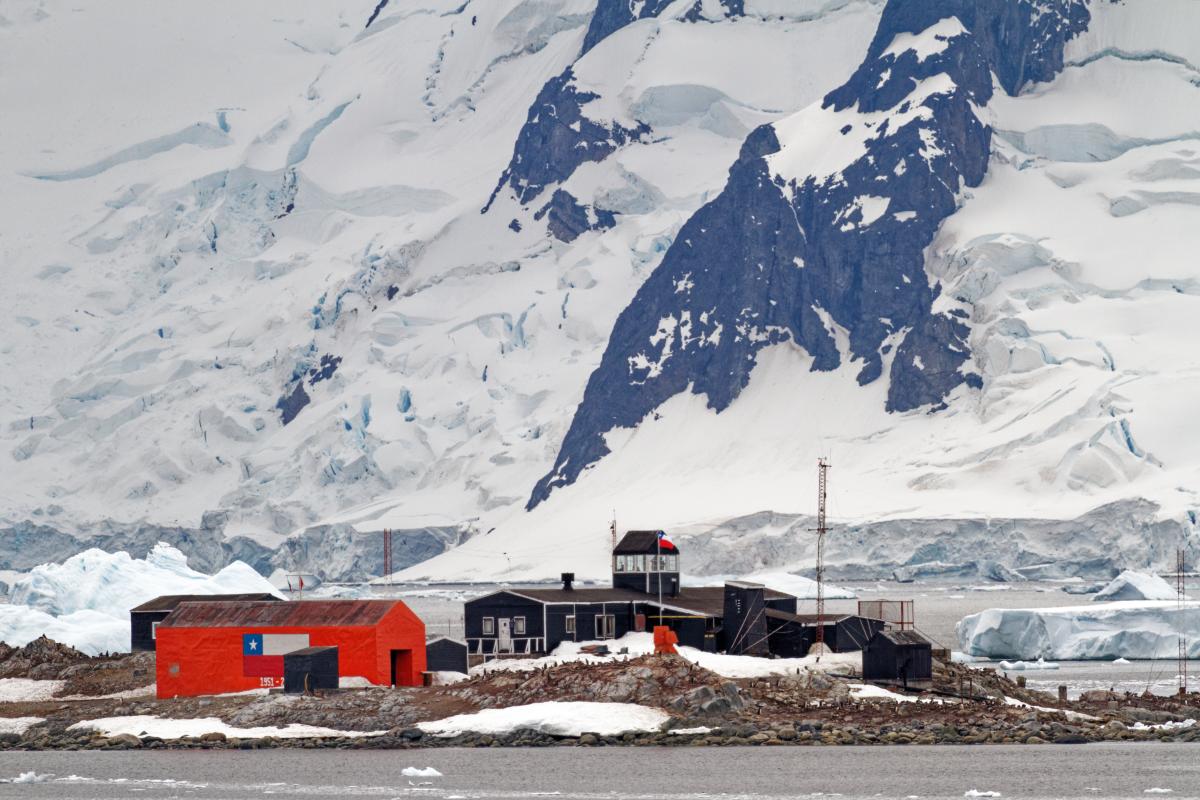 Small research station with black and red buildings sits on rocky, snow-covered terrain with icy mountains in the background.
