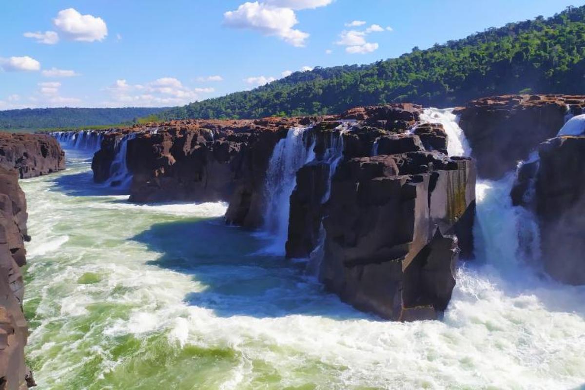 Wide river cascading over dark rocky cliffs, forming multiple waterfalls with lush green forest in the background under a blue sky with scattered clouds.