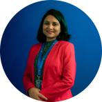 Sulagna Mishra in a red jacket standing in front of a blue background.