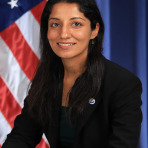 A person with long dark hair, wearing a black blazer with a small pin, poses in front of a United States flag.