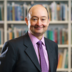 A person in a suit and tie stands in front of a bookshelf filled with books.