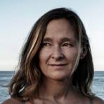 A woman with shoulder-length hair stands in front of the ocean, looking slightly to her right. The sky is clear and the water is calm in the background.