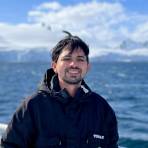 A man in a black jacket stands outdoors by a body of water with snowy mountains in the background on a sunny day.