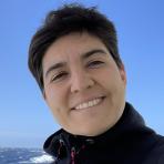 Person smiling outdoors with the ocean and a clear blue sky in the background.
