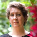 A woman with short wavy hair and blue earrings stands outdoors, facing the camera with a neutral expression. Green foliage and red flowers are in the background.