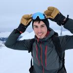 A person in winter gear stands on a snowy mountain, holding ski goggles above their head and smiling at the camera.