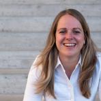 A woman with long blonde hair wearing a white shirt is smiling while sitting in front of a concrete staircase.