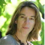 A woman with shoulder-length light brown hair is outdoors, wearing a light sweater and a necklace, with green foliage in the blurred background.