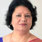 A woman with short, curly black hair, wearing earrings, a gold chain, a bindi on her forehead, and a saree with a pink border, poses against a plain light background.