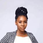 Woman with braided hair styled in a bun, wearing a houndstooth-patterned blazer and a white top, poses in front of a plain light background.