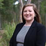 A woman with long brown hair, wearing a black blazer and patterned dress, stands outdoors in front of green foliage, smiling at the camera.
