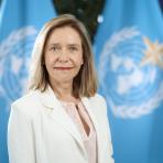 A woman in a white blazer stands in front of two blue flags featuring the United Nations emblem.