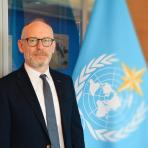 A man in a suit and glasses standing next to a blue World Meteorological Organization flag.