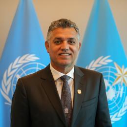 A person in a suit stands in front of two blue flags with the United Nations emblem.