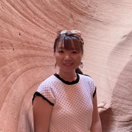 A person wearing a white shirt and glasses on their head stands in front of a curved, textured pink rock formation.