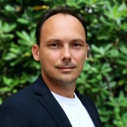 A man with short brown hair wearing a dark blazer and white shirt stands in front of green foliage, looking at the camera.