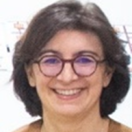 A person with short dark hair and glasses is smiling, wearing a brown top, in front of a white background with blurred details.