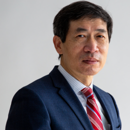 A man in a navy suit and red striped tie stands against a plain light background, looking at the camera with a neutral expression.