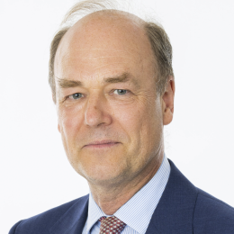 A middle-aged man with thinning light hair, wearing a blue suit, light blue shirt, and patterned tie, poses against a white background.