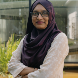 A woman wearing glasses and a dark purple hijab, standing outdoors with her arms crossed, in front of a modern glass building and greenery.