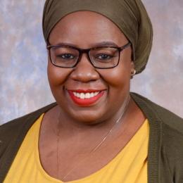 A woman wearing glasses, a green headscarf, and a yellow top smiles at the camera against a neutral, mottled background.