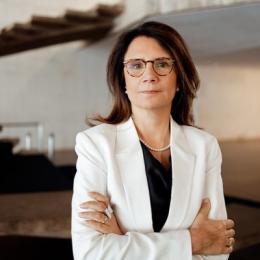 A woman with glasses and shoulder-length brown hair stands with arms crossed, wearing a white blazer and black top, in an indoor setting with modern architecture.