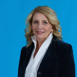 A woman with blonde hair wearing a dark blazer and white blouse stands against a solid blue background, looking at the camera.