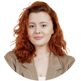 A woman with wavy red hair, wearing a tan blazer over a white top, poses against a plain white background.