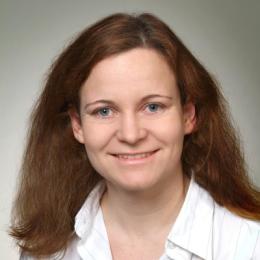 A woman with light skin, blue eyes, and shoulder-length brown hair is smiling at the camera, wearing a white collared shirt against a neutral background.