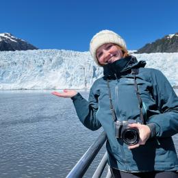 A person in a winter jacket and hat stands on a boat, smiling and gesturing toward a glacier and snowy mountains under a clear blue sky.