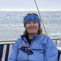 A woman in a blue jacket and headband sits on a boat with icy water and a glacier in the background.