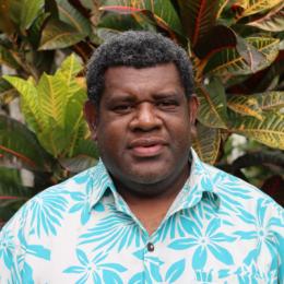 A man with short curly hair wearing a blue and white floral shirt stands in front of large, colorful tropical plants.