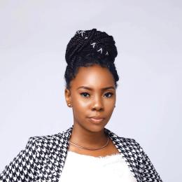 Woman with braided hair styled in a bun, wearing a houndstooth-patterned blazer and a white top, poses in front of a plain light background.