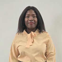 A woman with straight black hair wearing a light beige blouse with a bow stands against a plain light background, looking at the camera.