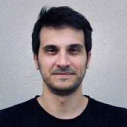 A man with short dark hair and light facial hair is wearing a black shirt and standing in front of a plain, light-colored wall.