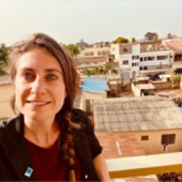 A woman with long brown hair in a braid stands on a balcony, smiling at the camera, with buildings and a clear sky in the background.