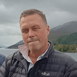 A man in a black Rab jacket stands outdoors near a body of water with mountains and trees in the background under a cloudy sky.