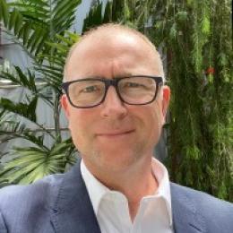 A man with glasses in a blue suit and white shirt stands outdoors in front of green plants, looking at the camera.