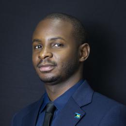 A man in a navy blue suit and tie poses against a dark background, wearing a lapel pin with a blue, yellow, and red flag.