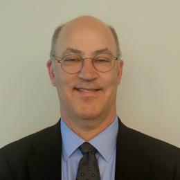 A middle-aged man with glasses, balding head, and a suit with a blue shirt and patterned tie, smiling in front of a plain light-colored background.