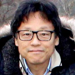 A person with short black hair and glasses is wearing a jacket with a fur-lined hood, standing outdoors.